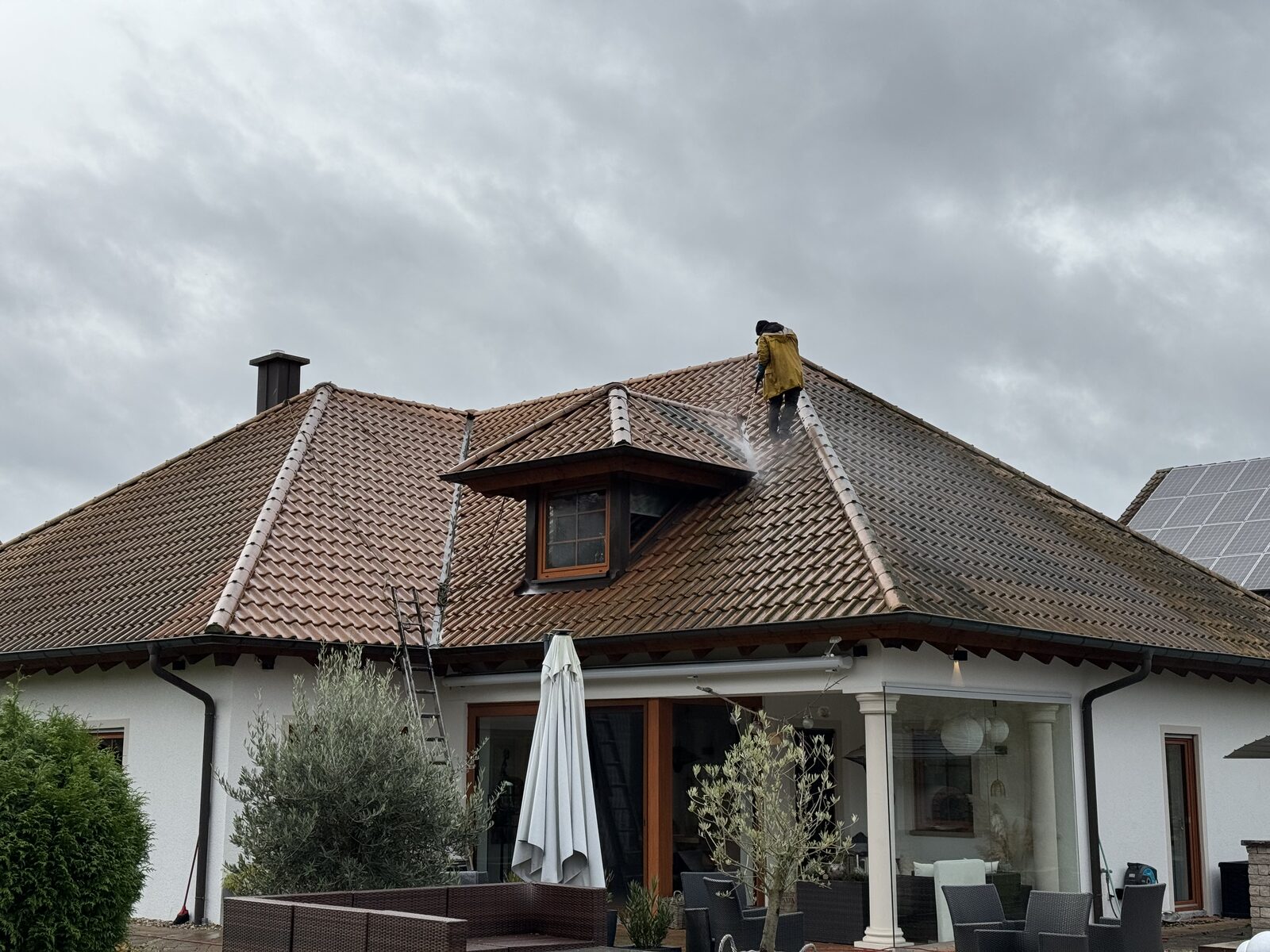 Weißes Einfamilienhaus während der Dachreinigung: Mitarbeiter im gelben Jacket mit Hochdruckreiniger auf dem Walmdach, Leiter und Terrassenmöbel